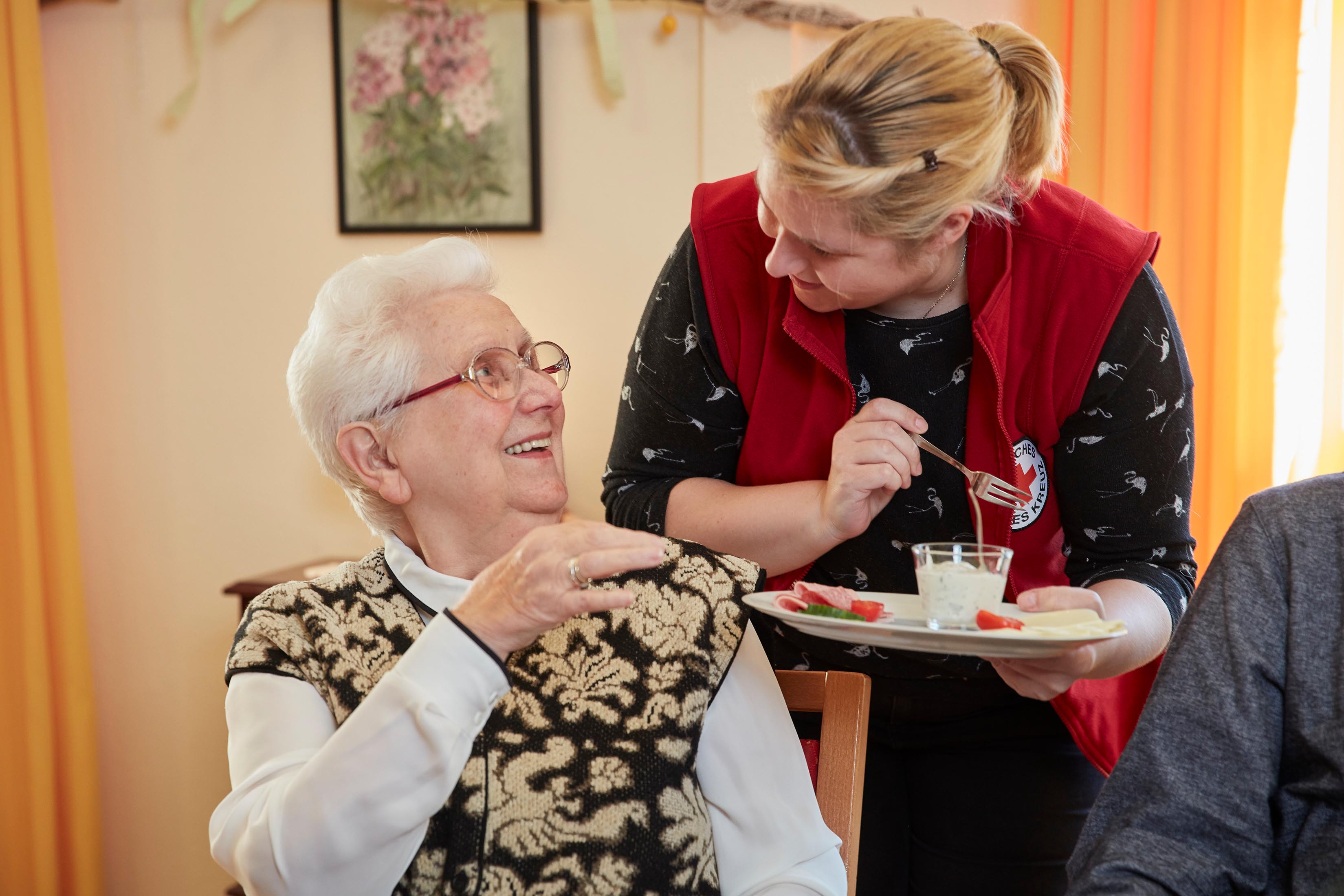 DRK-Mitarbeiterin reicht einer Seniorin einen Teller mit Essen, beide lachen