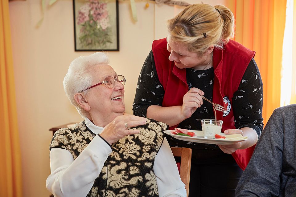 DRK-Mitarbeiterin reicht einer Seniorin einen Teller mit Essen, beide lachen